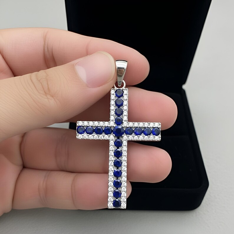 Pu&ograve; includere: Ciondolo a forma di croce in argento con un alone di piccole pietre bianche e pietre blu pi&ugrave; grandi. La croce &egrave; tenuta davanti a un portagioie nero.