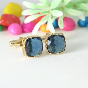 May include: A pair of gold-toned cufflinks with square, blue-grey gemstone centers. The cufflinks have a textured border and a classic post closure. Colorful decorative stones are in the background.