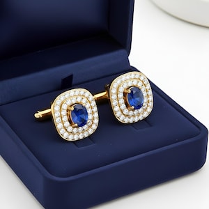 Peut inclure: Paire de boutons de manchette dorés de forme carrée. Chaque bouton de manchette présente une pierre précieuse bleue ovale centrale, entourée de petits diamants transparents. Présentés dans un écrin en velours bleu foncé.