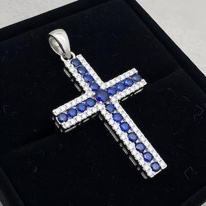 Pu&ograve; includere: Ciondolo a forma di croce in argento, ornato da pietre preziose blu e bianche. La croce presenta una fila centrale di pietre blu, bordata da piccole pietre bianche. Il ciondolo &egrave; presentato su uno sfondo nero.