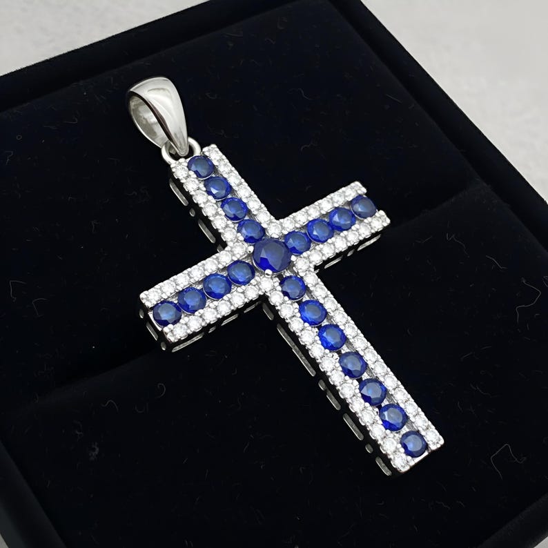 Pu&ograve; includere: Ciondolo a forma di croce in argento, ornato da pietre preziose blu e bianche. La croce presenta una fila centrale di pietre blu, bordata da piccole pietre bianche. Il ciondolo &egrave; presentato su uno sfondo nero.