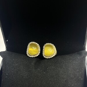 Op de afbeelding: Een paar gele edelsteen oorknopjes met een goudkleurige zetting en een halo van kleine, heldere kristallen. De oorbellen worden gepresenteerd in een zwarte sieradendoos.