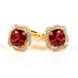 May include: A pair of gold-toned cufflinks with square, deep red gemstones at the center. Each cufflink is surrounded by a halo of small, clear gemstones. The cufflinks have a classic design, suitable for formal wear.