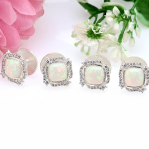 May include: Four silver stud earrings with square opal centers and a halo of small, clear gemstones. The earrings are displayed on a white surface, with pink and white flowers in the background.