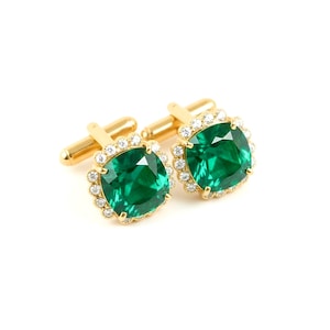 May include: A pair of gold cufflinks featuring large, square-cut emerald green gemstones. Each gemstone is surrounded by a halo of small, clear crystals. The cufflinks have a classic design with a T-bar closure.