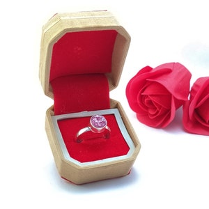 Peut inclure: Une bague avec une pierre rose dans une petite boîte en bois avec un intérieur en velours rouge. La boîte est ouverte et la bague est à l'intérieur. Deux roses rouges sont en arrière-plan.