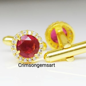 May include: A pair of gold cufflinks with a red gemstone surrounded by a halo of white gemstones. The cufflinks are connected by a gold bar.