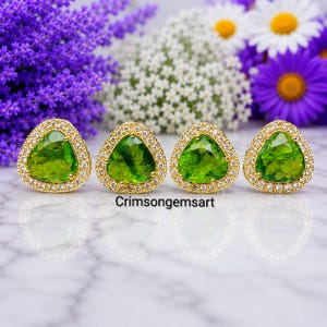 Op de afbeelding: Vier driehoekige oorbellen met levendige groene gefacetteerde edelstenen in goudkleurige metalen monturen, omrand door kleine heldere steentjes. De tekst "Crimsongemsart" is in het zwart onder de vier oorbellen gedrukt.