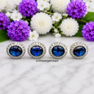 May include: Four silver-toned stud earrings with oval, deep blue center stones. Each earring is surrounded by a halo of small, clear crystals. The earrings are displayed on a white marble surface, with purple and white flowers in the background.
