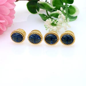May include: Four gold-toned cufflinks with oval, dark blue gemstone centers. The cufflinks have a detailed, textured design around the gemstones. The image is set against a white background with floral accents, suggesting a jewelry or accessory theme.