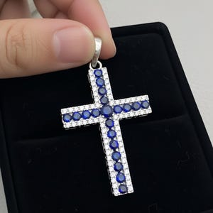 Pu&ograve; includere: Ciondolo a forma di croce color argento con una fila di pietre preziose blu al centro e pietre preziose bianche sui bordi. Il ciondolo &egrave; mostrato su uno sfondo nero.