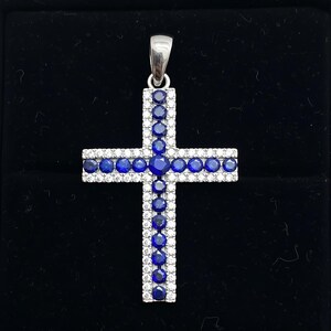 Pu&ograve; includere: Un ciondolo a forma di croce in argento con un'ansa. La croce &egrave; adornata con file alternate di pietre rotonde e chiare e pietre rotonde blu scuro. Il ciondolo &egrave; posto su uno sfondo nero.