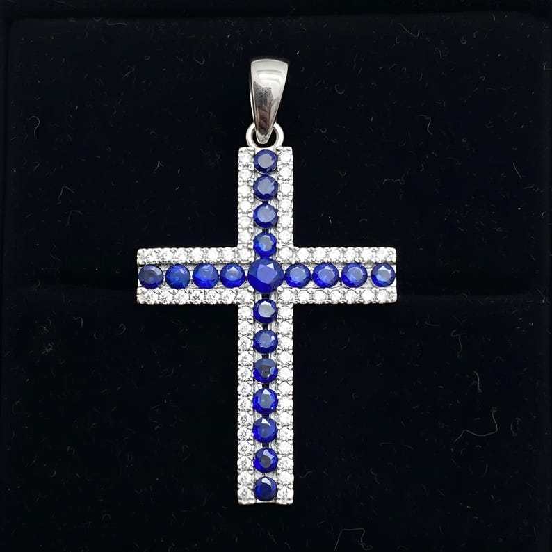 Pu&ograve; includere: Un ciondolo a forma di croce in argento con un'ansa. La croce &egrave; adornata con file alternate di pietre rotonde e chiare e pietre rotonde blu scuro. Il ciondolo &egrave; posto su uno sfondo nero.