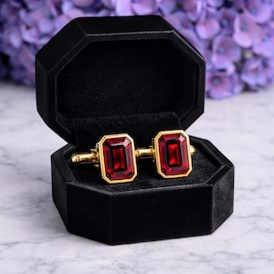 May include: A pair of gold-toned cufflinks with rectangular, deep red gemstone centers. The cufflinks are displayed in an open, black velvet octagonal box, set against a blurred background of purple flowers and a marble surface.