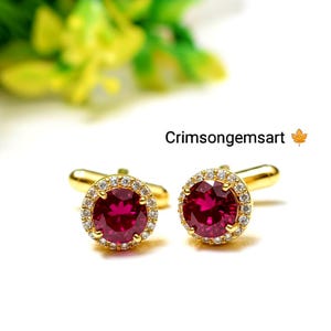 May include: Two gold cufflinks, each featuring a large, round, deep red gemstone encircled by smaller, clear stones. The cufflinks are set against a white backdrop, with the text "Crimsongemsart".