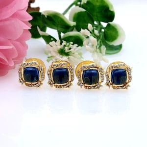 May include: Four gold stud earrings, each featuring a square, dark blue centre stone. The stones are framed by small, clear crystals. The earrings are presented on a white surface, with pink and green floral decorations in the background.