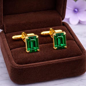 Puede incluir: Un par de gemelos dorados con piedras preciosas rectangulares de color verde esmeralda, presentados en una caja de terciopelo marrón. Un accesorio elegante para ocasiones formales, que añade un toque de sofisticación.