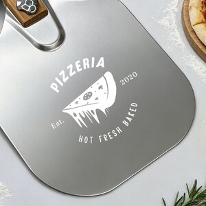 Könnte beinhalten: Ein Pizzaschieber aus Edelstahl mit Holzgriff und einer Lederschlaufe zum Aufhängen. Die Metalloberfläche zeigt eine weiße Grafik eines Pizzastücks mit dem kreisförmigen Text "PIZZERIA", "Est. 2020" und "HOT FRESH BAKED".