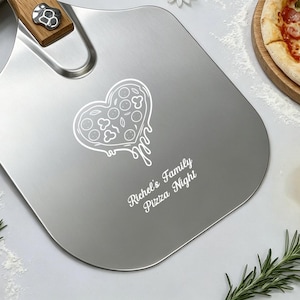 Könnte beinhalten: Ein Pizzaschieber aus Edelstahl mit Holzgriff, der eine lasergravierte herzförmige Pizza-Grafik und den Text "Richel's Family Pizza Night" auf der Klinge aufweist. Das Werkzeug hat eine Metallniete, die den Griff mit der flachen Oberfläche verbindet.