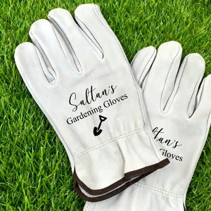 May include: A pair of white leather gardening gloves with brown trim, resting on green grass. The gloves are personalized with the text "Saltan's Gardening Gloves" and a small shovel graphic.
