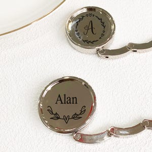Könnte beinhalten: Zwei silberne Handtaschenaufhänger mit kreisförmigen Oberteilen. Einer ist mit dem Buchstaben "A" in einem Kranzdesign graviert, der andere mit dem Namen "Alan" und einem ähnlichen Kranzdesign. Die Aufhänger haben ein faltbares Design für einfache Aufbewahrung.