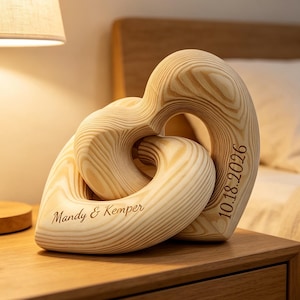 May include: A light-colored wooden sculpture consisting of two interlocking heart shapes. The wood features a visible grain pattern. One heart is engraved with the text Mandy & Kemper, and the other heart is engraved with the date 10.18.2026.
