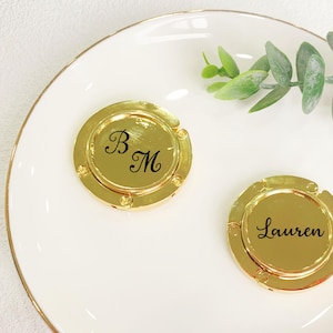 Puede incluir: Dos soportes dorados para teléfono, uno con "B M" y otro con "Lauren" en escritura negra. Se encuentran sobre un plato blanco con borde dorado, acompañados de una ramita verde.