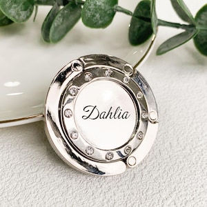 May include: A silver-coloured purse hook with a white centre, adorned with small, clear crystals. The word "Dahlia" is written in black script across the centre. The hook is circular with a hinged design.