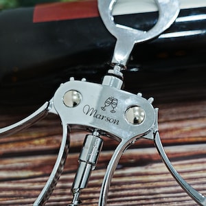 May include: A silver-toned metal wing corkscrew featuring a central engraving of two clinking wine glasses and the name Marson. The tool has two side levers, a central worm, and a circular top handle, all with a polished, reflective finish.