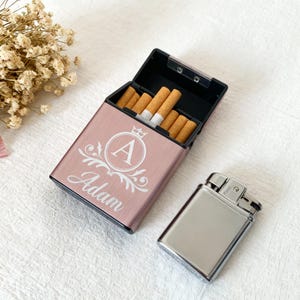 May include: A rose gold cigarette case with the name "Adam" and the letter "A" in a decorative design. The case is open, revealing cigarettes. A silver lighter sits beside the case.