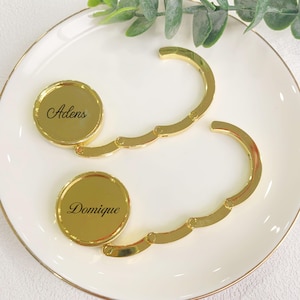Puede incluir: Dos colgadores de bolso dorados con bases circulares y brazos curvos. Las bases están inscritas con los nombres "Adens" y "Domique" en escritura negra. Los colgadores se exhiben en un plato blanco con borde dorado, con follaje verde en el fondo.