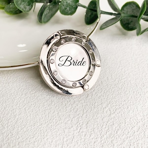 Puede incluir: Un gancho plateado para bolso con diseño circular. El centro presenta la palabra "Bride" en elegante escritura, rodeada de pequeños cristales brillantes. El gancho descansa sobre una superficie blanca con borde dorado.