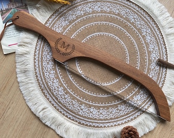 May include: A wooden cake server with a clear blade and a brown leather loop. The server features an engraved initial "M" within a laurel wreath. The server is on a round, fringed placemat.