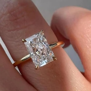 May include: A gold ring with a large, rectangular-cut diamond. The clear diamond sparkles, set on a slender, yellow gold band. The ring is displayed on a finger, showcasing the elegant design and the brilliance of the gemstone.