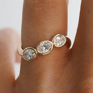 Può includere: Un anello d'oro con tre diamanti ovali disposti in fila. I diamanti trasparenti sono incastonati in una fascia semplice. L'anello è mostrato su un dito, evidenziando il suo design elegante. Il design dell'anello è delicato e raffinato.