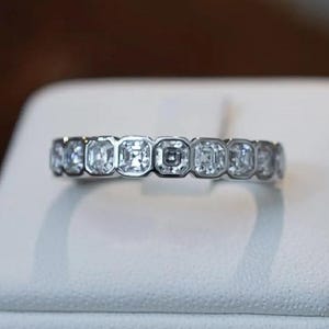 May include: A silver ring with a series of square-cut, clear gemstones set around the band. The ring is displayed on a white surface, with a blurred background of brown and white.