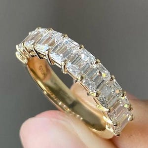 May include: A gold band ring featuring a series of emerald-cut, clear gemstones. The ring's band is a polished gold color, and the gemstones are set closely together, creating a continuous line of sparkle. The ring is held up against a neutral background.