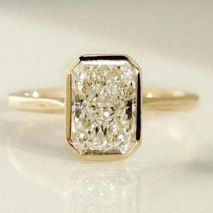 Op de afbeelding: Een gouden ring met een rechthoekig geslepen diamant. De diamant is gezet in een bezel en de ring wordt getoond op een reflecterend oppervlak. De facetten van de diamant reflecteren het licht, waardoor een sprankelend effect ontstaat.