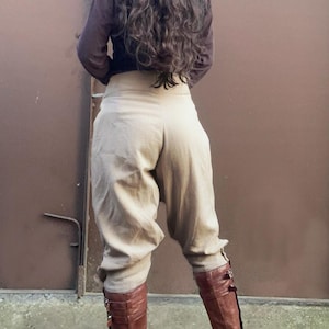 May include: Tan high-waisted pants with a gathered, voluminous design. The pants are paired with brown leather knee-high boots with buckle details. A long-sleeved brown shirt completes the outfit, creating a cohesive, earthy aesthetic.
