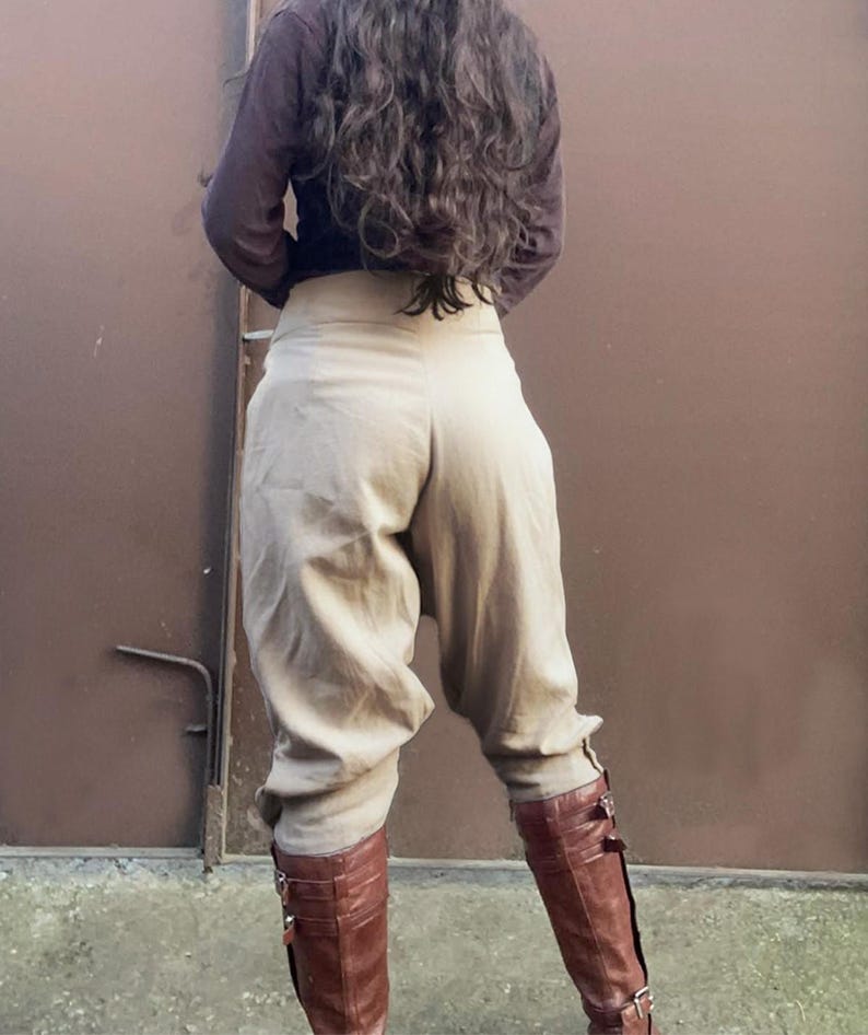 May include: Tan high-waisted pants with a gathered, voluminous design. The pants are paired with brown leather knee-high boots with buckle details. A long-sleeved brown shirt completes the outfit, creating a cohesive, earthy aesthetic.