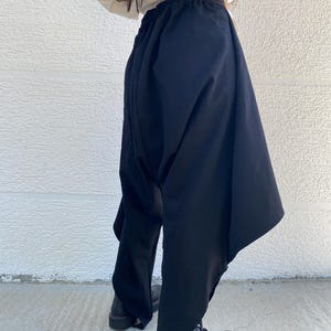 May include: Black drop-crotch trousers featuring an elasticized waistband and a voluminous, asymmetrical silhouette that tapers toward the ankles. The pants are constructed with deep, draped fabric panels on the sides, creating a distinctively loose and flowing shape.