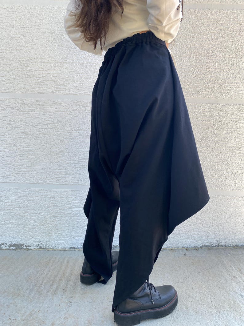 May include: Black drop-crotch trousers featuring an elasticized waistband and a voluminous, asymmetrical silhouette that tapers toward the ankles. The pants are constructed with deep, draped fabric panels on the sides, creating a distinctively loose and flowing shape.
