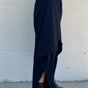 May include: Black trousers featuring an asymmetrical layered design with a high-low hemline and side slits, paired with black lace-up platform boots. The trousers have a fitted waistband and a flowing silhouette that extends down to the ankles.