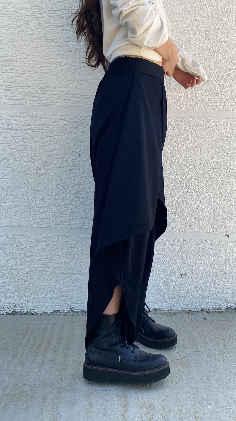 May include: Black trousers featuring an asymmetrical layered design with a high-low hemline and side slits, paired with black lace-up platform boots. The trousers have a fitted waistband and a flowing silhouette that extends down to the ankles.