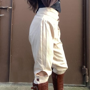 May include: Cream-colored, high-waisted pants with a unique design, featuring button details and cut-out accents near the ankles. Paired with brown leather boots. The pants have a pleated front and a tailored fit.
