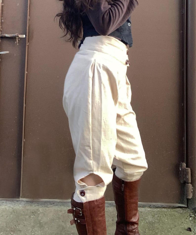 May include: Cream-colored, high-waisted pants with a unique design, featuring button details and cut-out accents near the ankles. Paired with brown leather boots. The pants have a pleated front and a tailored fit.