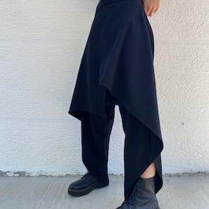 May include: Black trousers featuring an asymmetrical wrap-style overlay panel that drapes over the legs. The garment has a high waistband and a structured silhouette, paired with black lace-up platform boots and a cream-colored long-sleeve top.