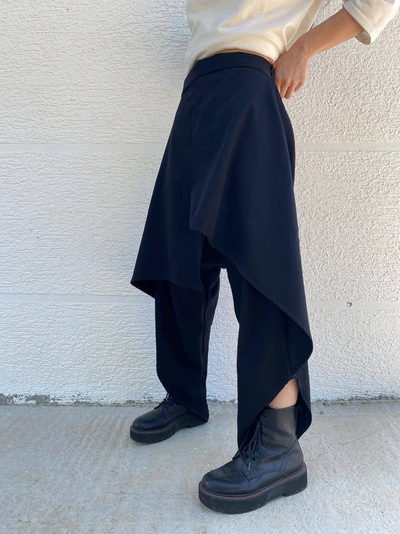 May include: Black trousers featuring an asymmetrical wrap-style overlay panel that drapes over the legs. The garment has a high waistband and a structured silhouette, paired with black lace-up platform boots and a cream-colored long-sleeve top.