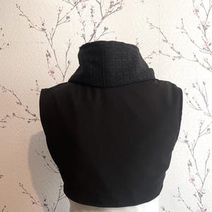 Pode incluir: Uma pe&ccedil;a de vestu&aacute;rio preta sem mangas com uma gola texturizada cinza escuro. A gola &eacute; um estilo de gola alta. A pe&ccedil;a &eacute; feita de um tecido liso e fosco. O fundo &eacute; um papel de parede floral.