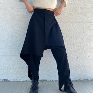 May include: Black trousers featuring an asymmetrical wrap-style skirt overlay that drapes down the front. The garment has a high waistband and wide-leg cut, paired with black lace-up boots. The fabric appears solid and opaque in texture.